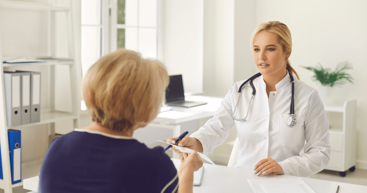 A female doctor in a white lab coat sitting at a bright desk, handing a document to a blonde female patient seated across from her.