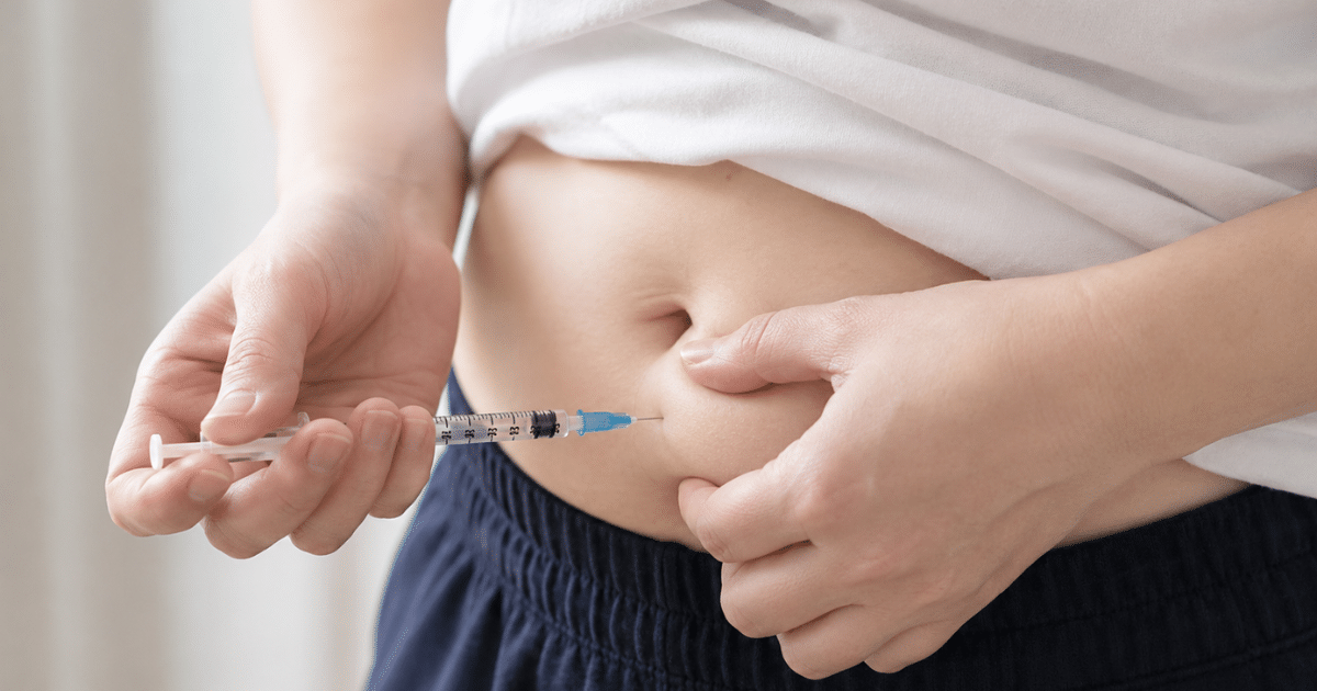 A close-up shot showing a person pinching a fold of skin near their belly button with one hand while using a clear syringe with the other hand to inject medication directly into the subcutaneous fat of their abdomen.