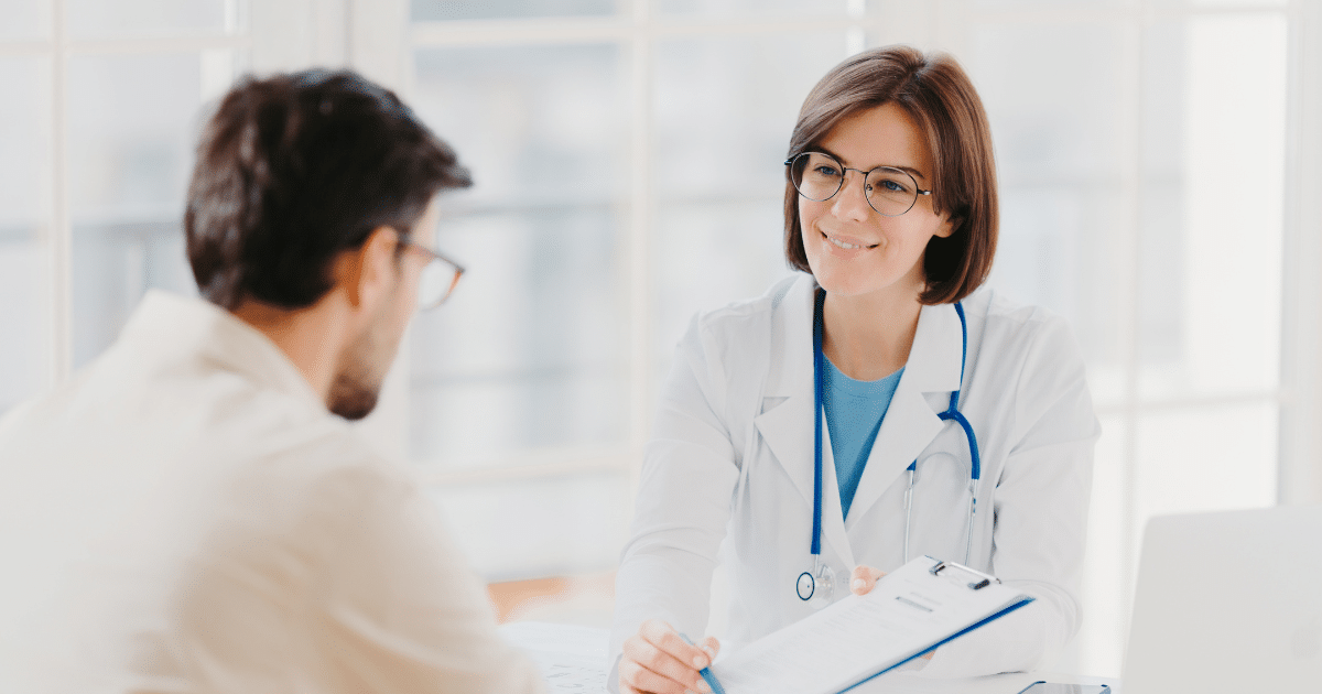 A female doctor with short brown hair and glasses smiling warmly while presenting a medical document on a clipboard to a patient across her desk.