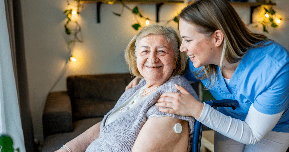 An smiling elderly woman wearing a grey knit cardigan sits near a window, with a round white continuous glucose monitor attached to her upper arm. Behind her, a smiling caregiver in blue scrubs gently places a hand on her shoulder.