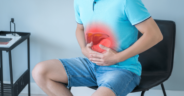 A man in a blue polo shirt and denim shorts sitting on a chair, holding his stomach with a red glowing area highlighting liver and stomach pain.