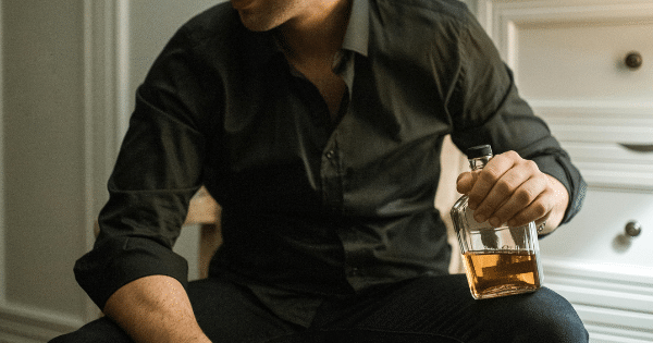 man wearing an open black button-down shirt sitting down while holding a glass bottle filled with amber liquor.