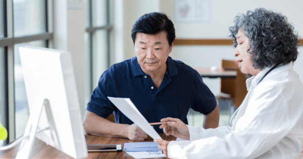 A doctor sitting across from a male patient at a desk, reviewing medical documents together.