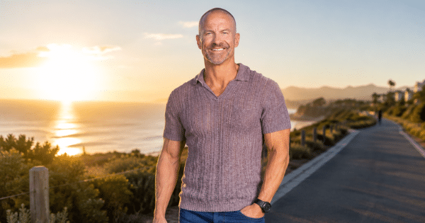A fit, smiling man in a knit short-sleeve shirt standing on an outdoor coastal pathway during sunset.