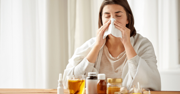A woman wrapped in a blanket blowing her nose with a tissue behind a table of home remedies and medicines.