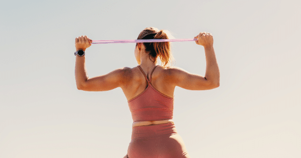 A view from behind of a athletic woman stretching a pink resistance band above her head outdoors.