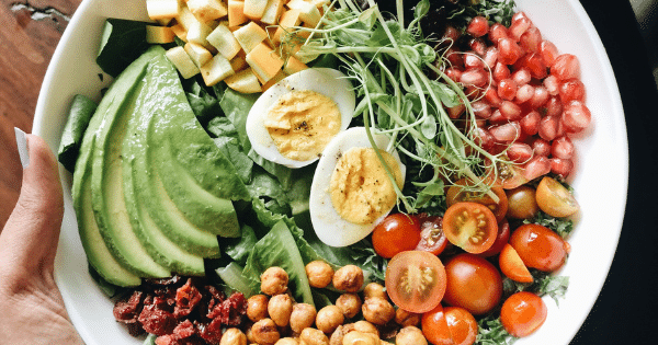 A healthy meal bowl filled with avocado, halved hard-boiled eggs, chickpeas, tomatoes, pomegranate seeds, and greens.