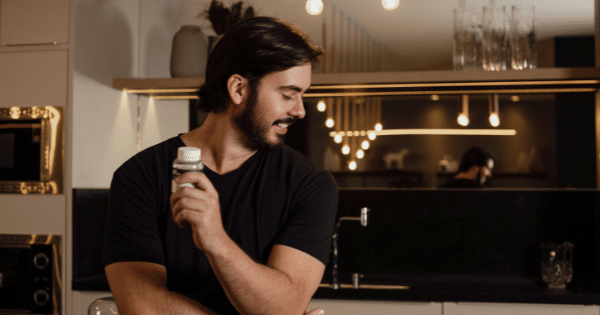 A smiling man in a dark t-shirt holding a small supplement bottle in a modern kitchen setting.
