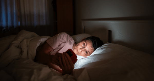 A middle-aged woman lying in bed in a dark room, holding a glowing smartphone near her face.