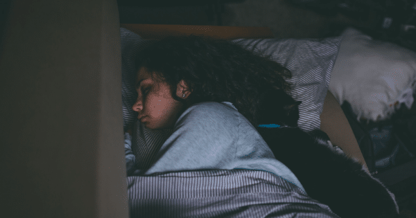 A woman sleeping on a couch under a striped blanket with a black cat curled up next to her head.