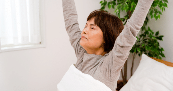 An older woman sitting in bed with her eyes closed, stretching her arms overhead as she wakes up in a bright room.