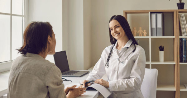 A smiling female doctor wearing a white lab coat sitting at a desk with a laptop, speaking with a patient seen from behind.