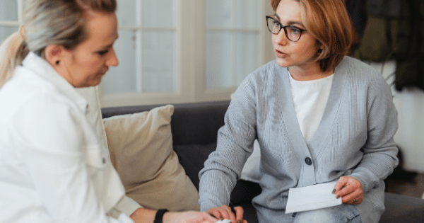 A female healthcare worker wearing glasses and a cardigan sitting on a couch and talking intently with a patient.