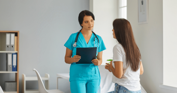 A female nurse wearing bright blue scrubs and a stethoscope holding a clipboard while talking to a patient standing in a medical office.