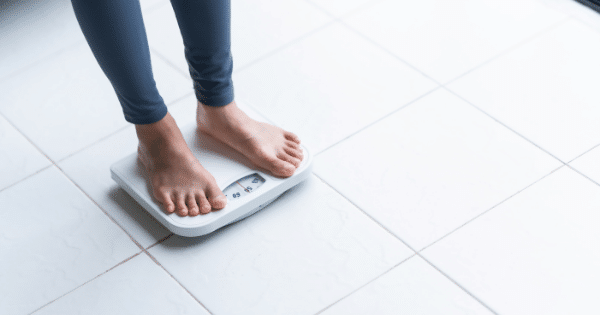 Close-up of a person's bare feet standing on a white analog bathroom scale on a tiled floor.