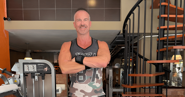 A fit man standing with folded arms in a gym near a workout machine and a wooden spiral staircase.