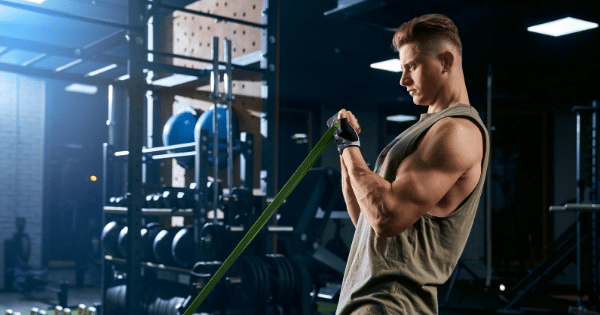 A man in a workout tank top performing a bicep curl exercise using a green resistance band inside a gym.