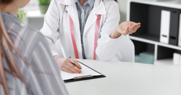 Close-up of a doctor wearing a white coat with a pink measuring tape around her neck, gesturing open-handed while consulting with a patient at a desk.