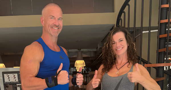 A muscular man in a blue tank top and a woman in a grey tank top giving double thumbs-up inside a gym setting.