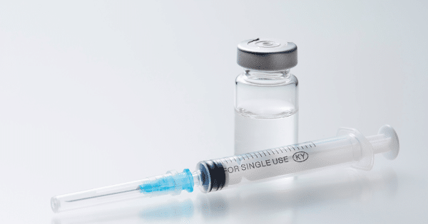 A clear glass medical vial with a silver cap resting next to a transparent plastic syringe with a needle cover.
