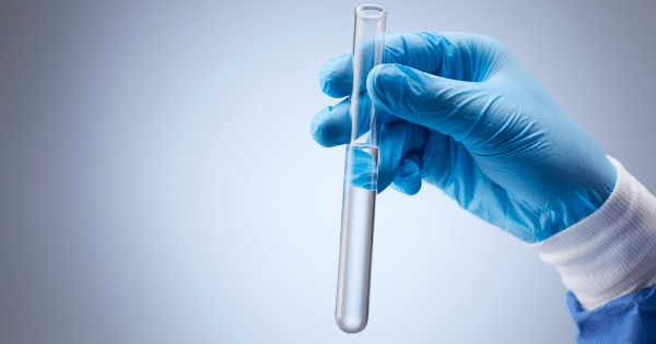 Hand wearing a blue gloved holding a test tube filled with clear liquid.