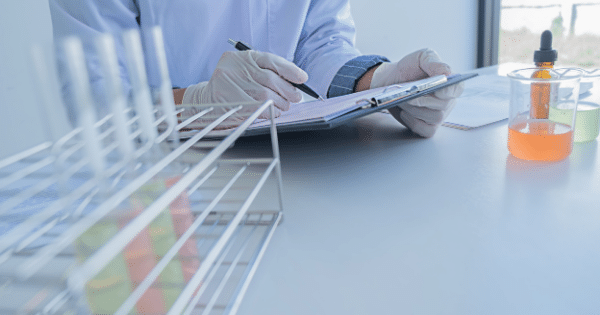 Lab technician wearing white gloves filling out documentation near beakers and test tubes.