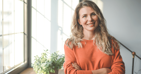 Blonde woman in an orange sweater posing indoors with folded arms near a window.