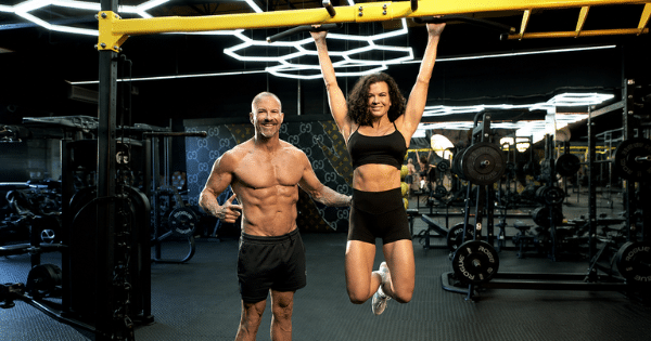 A fit woman hanging from a pull-up bar beside a shirtless, muscular man gesturing toward her in a dark gym.