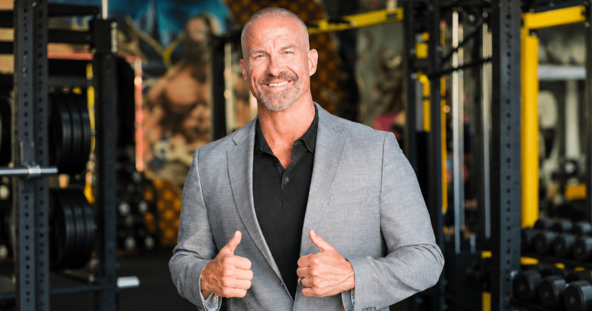 Smiling man in a grey blazer giving two thumbs up inside a gym setup.
