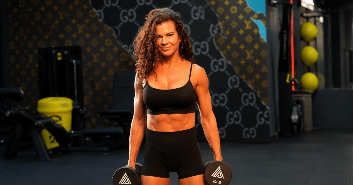 A athletic woman with curly brown hair wearing a black sports bra and shorts stands in a gym, holding dumbbells in both hands while facing the camera.