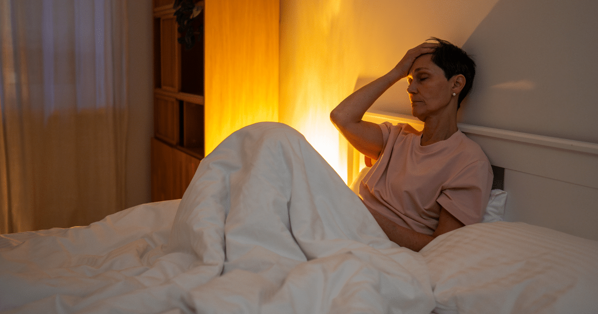 A middle-aged woman sitting up in bed under a blanket in a dimly lit room, holding her hand to her forehead with a tired expression.