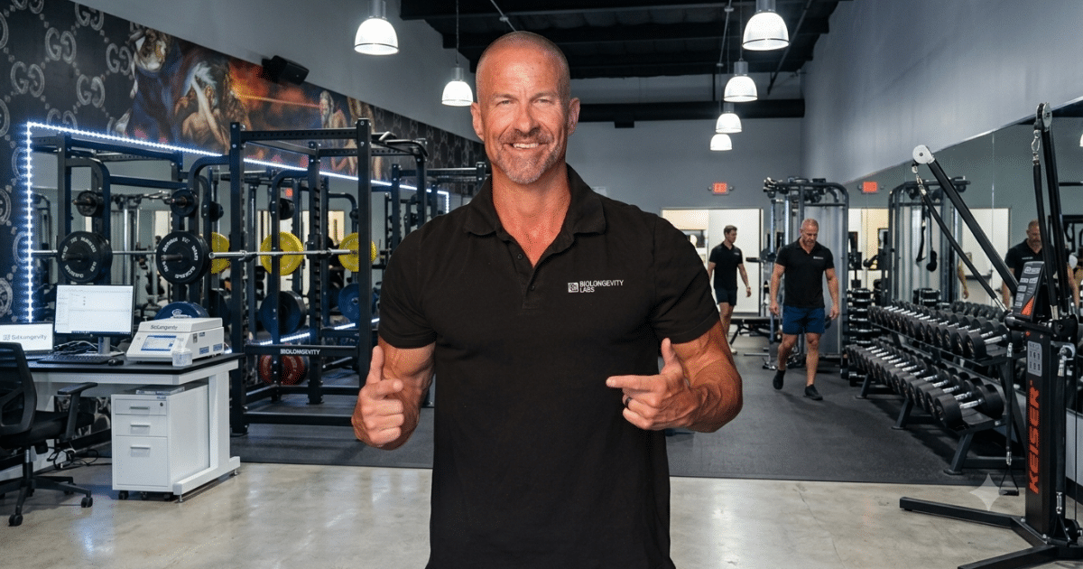 Man in a black polo shirt pointing towards himself, standing inside a gym facility with weight racks and exercise equipment in the background.