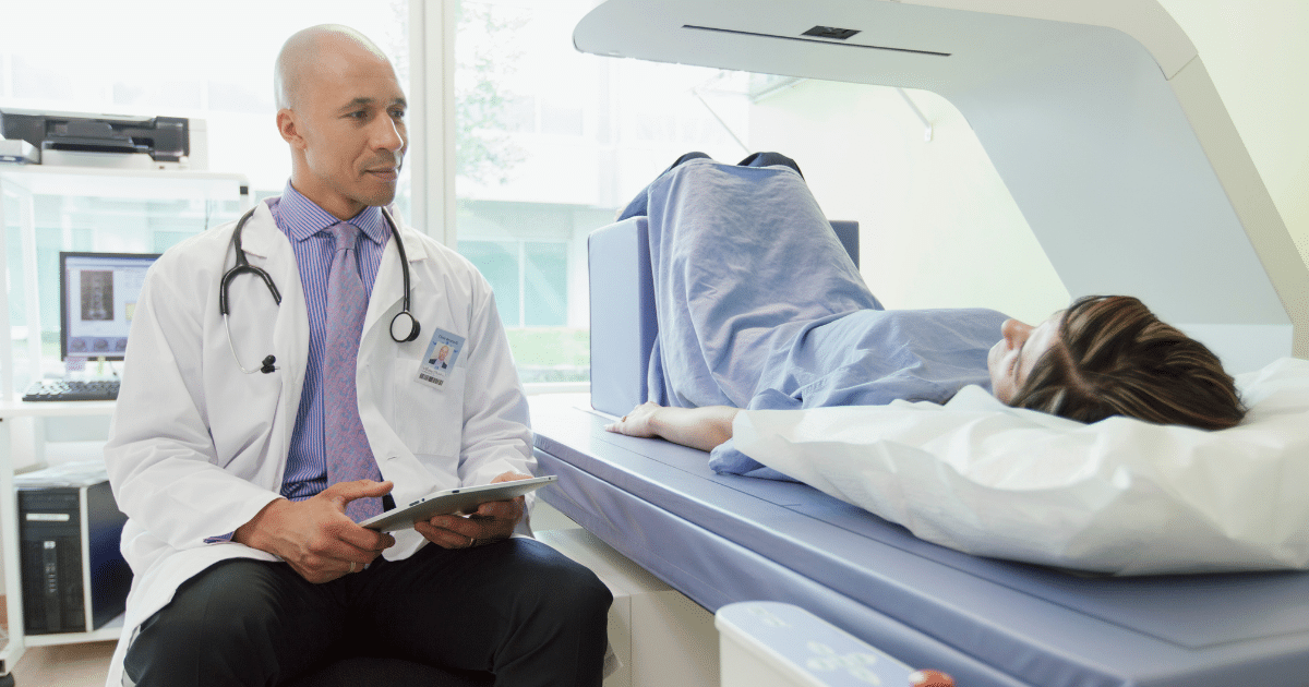 A male doctor in a white lab coat sitting next to a DXA scan machine holding a tablet while a female patient lies under the scanner.
