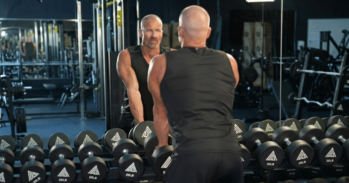 Rear view of a bald, muscular man in a black tank top resting his hands on a dumbbell rack, seen in reflection in a gym mirror.