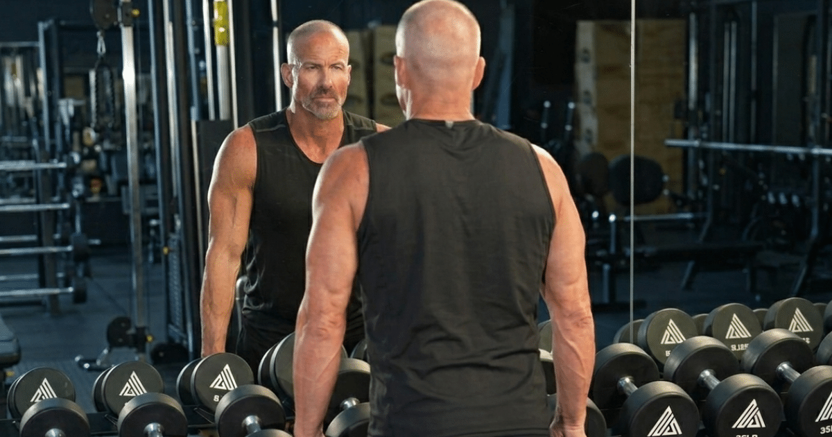 A fit, middle-aged man with a shaved head wearing a black tank top standing in a gym, facing away from the camera and looking into a mirror in front of a rack of dumbbells.