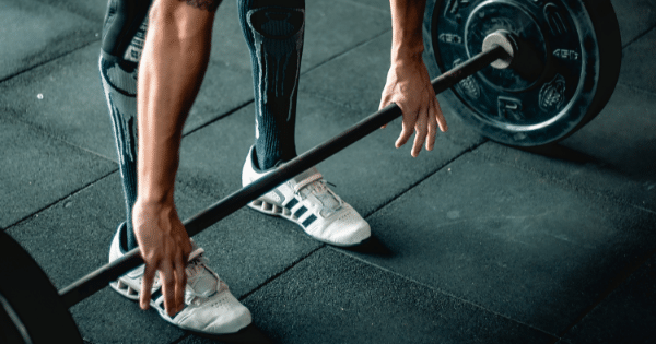 A low-angle close-up of a person in weightlifting shoes and high socks reaching down to grip a loaded barbell resting on the gym floor, preparing for a deadlift.