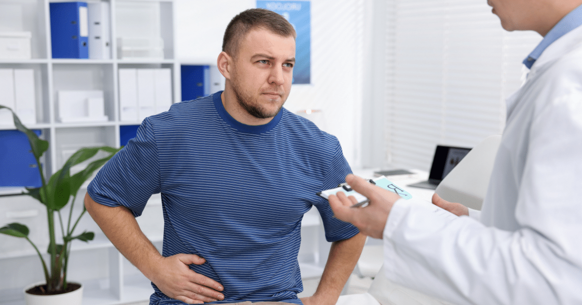 A patient seated on an examination table holds his side in discomfort while discussing symptoms with a doctor holding a clipboard.