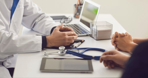 Doctor in a white coat seated at a desk across from a patient during a consultation, with a stethoscope and tablet on the table.