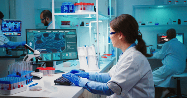 Female scientist wearing lab coat and blue gloves working on a computer displaying microscopic data inside a modern laboratory.