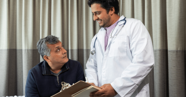 Male doctor in a white lab coat holding a clipboard and smiling while conversing with a seated male patient.