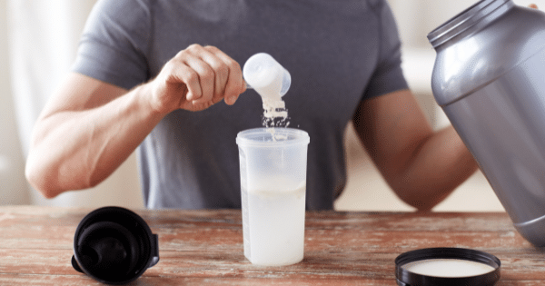 Close-up of a man pouring a scoop of white protein powder into a shaker bottle next to a large plastic tub.