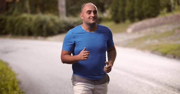 Man in a bright blue athletic shirt and shorts jogging outdoors along a paved road lined with trees.