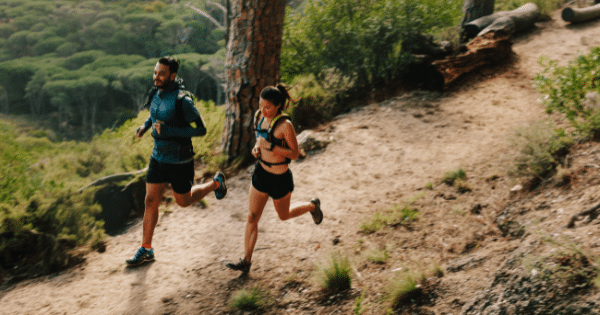 A man and a woman in athletic wear and hydration vests running along a dirt trail on a hillside surrounded by lush green trees.
