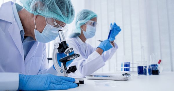 Two lab technicians in protective suits and face masks conducting research with a microscope and test tubes inside a laboratory.