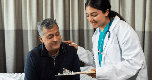 Smiling female doctor wearing a lab coat and stethoscope placing a supportive hand on a male patient's shoulder.