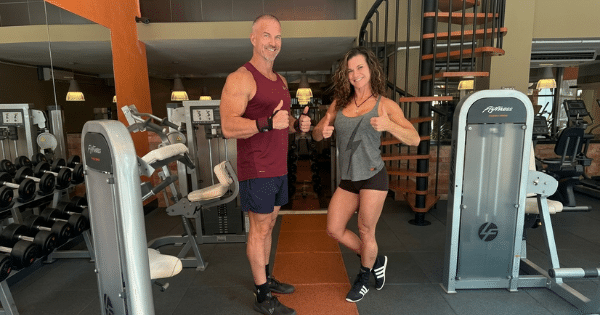 Athletic man and woman giving thumbs-up gestures while standing between weight machines in a gym with a spiral staircase in the background.