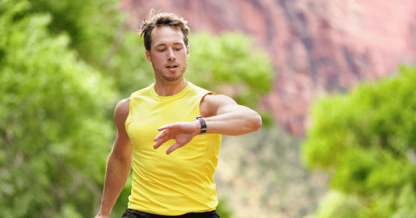 A young man in a sleeveless yellow athletic shirt checking his smartwatch while running outdoors against a blurred background of green foliage and red rock.
