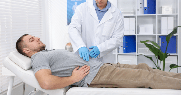A patient lies reclined on an examination table while a doctor wearing a white lab coat and blue gloves performs an abdominal examination.