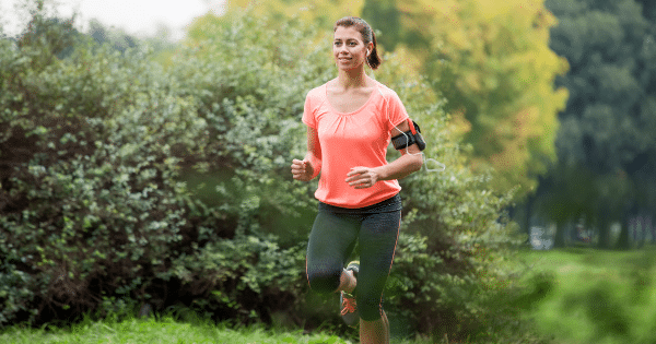 A smiling woman jogging along a tree-lined path in a park, wearing an orange workout shirt, black leggings, and an armband holding her phone with wired earphones.