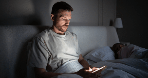 A man sits up in bed in a dark room illuminated only by his glowing smartphone screen while a partner sleeps nearby.